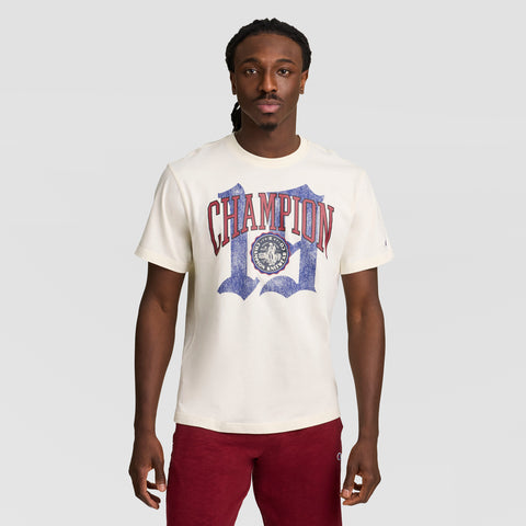 A man with long braided hair wears a cream Modern Retro Gothic '19 T-Shirt, C Logo by Champion in red and blue, paired with red pants. He stands against a plain light gray background.