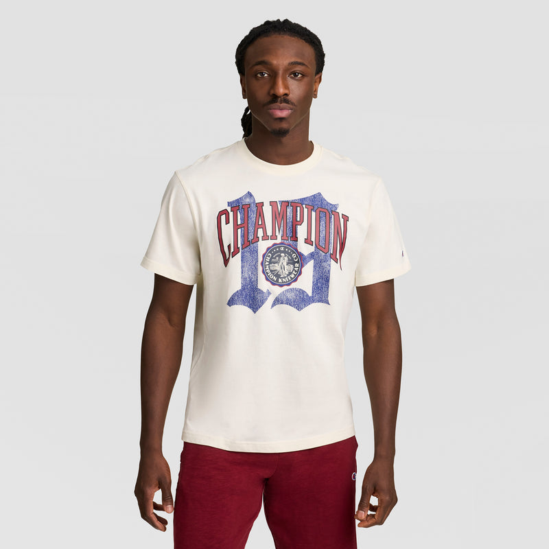 A man with long braided hair wears a cream Modern Retro Gothic '19 T-Shirt, C Logo by Champion in red and blue, paired with red pants. He stands against a plain light gray background.