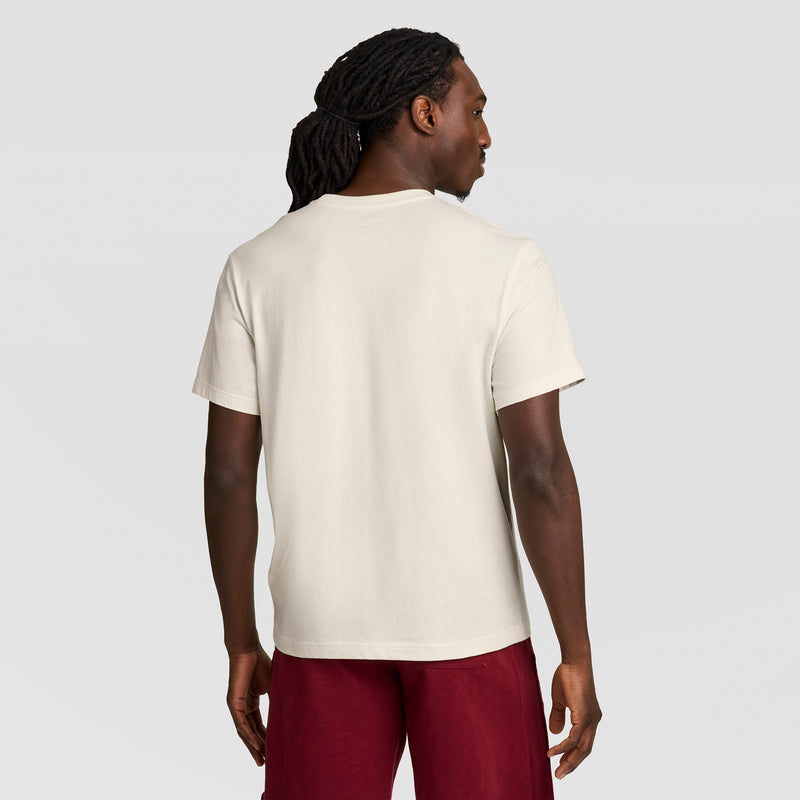 A man with long, dark, braided hair wears a Champion Modern Retro Gothic '19 T-Shirt with a C Logo and red shorts, standing with his back to the camera against a plain gray background.