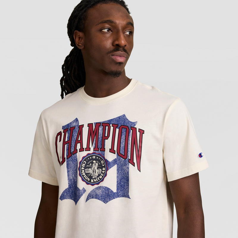 A man with long dark twisted hair wears a cream Modern Retro Gothic '19 T-Shirt by Champion, featuring bold red "CHAMPION" lettering and a blue C logo, as he looks slightly left against a plain light gray background.