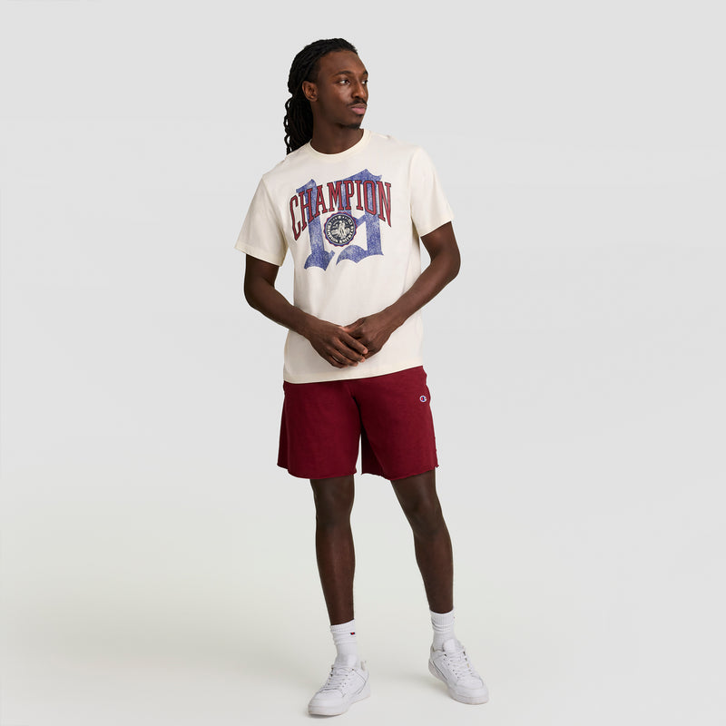 A person stands against a plain background, wearing a cream-colored Champion Modern Retro Gothic '19 T-Shirt with the C Logo, maroon shorts, white socks, and white sneakers. Their long hair is braided as they look to the side.