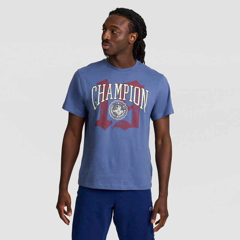 A man in a blue Champion Modern Retro Gothic '19 T-Shirt, featuring the C logo, and navy shorts stands against a plain light gray background, looking left with a neutral expression.