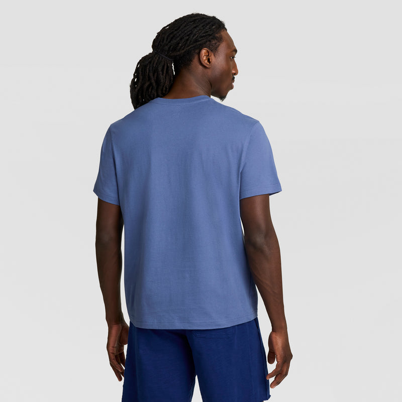 A man with long, dark, braided hair is seen from behind, wearing a blue Champion Modern Retro Gothic '19 T-Shirt with a C logo and blue shorts, standing against a plain, light gray background.