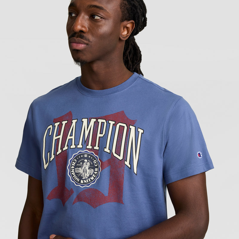 A man with long dark hair models the blue Champion Modern Retro Gothic '19 T-Shirt, featuring a bold C Logo graphic and crest, looking to the side against a light background—a standout men’s fashion tee.