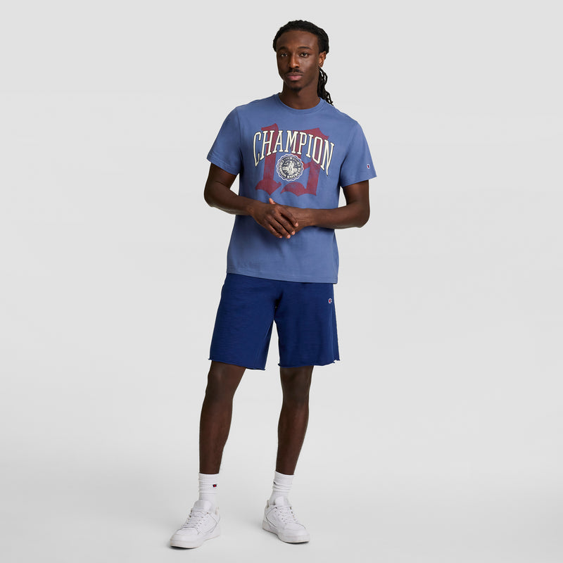 A man with long braids poses relaxedly against a plain background wearing a blue Champion Modern Retro Gothic '19 T-Shirt with C Logo, dark blue shorts, white socks, and white sneakers. One hand touches his fingers.