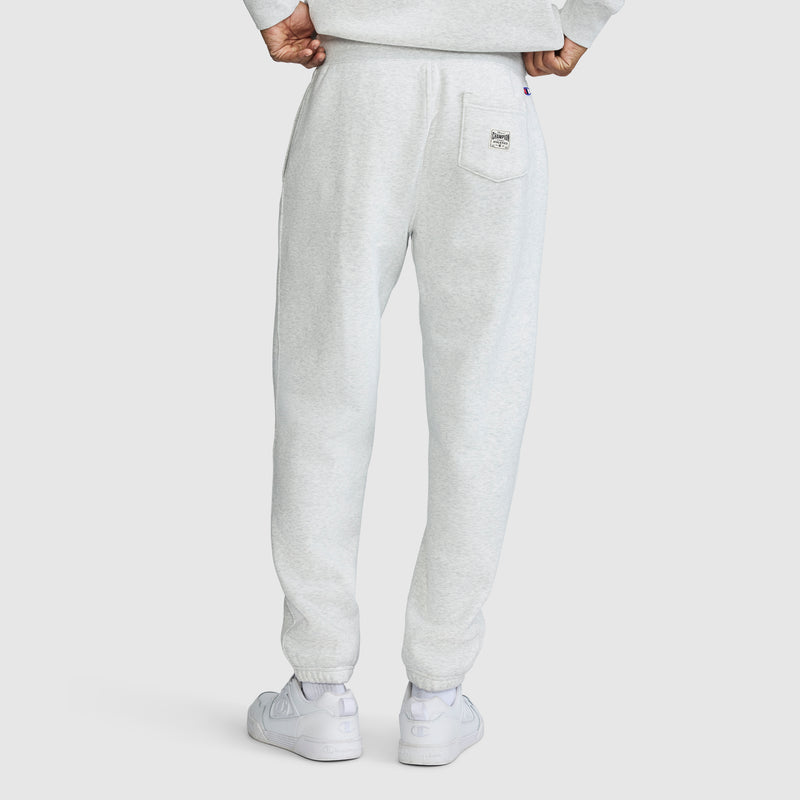 A person wearing Champion All in a Day's Work Sweatpants in light gray and white sneakers is shown from the back. The sweatpants feature a right-side back pocket, a signature detail of Champion men's styles. The person's hands are tucked into the waistband at the back.