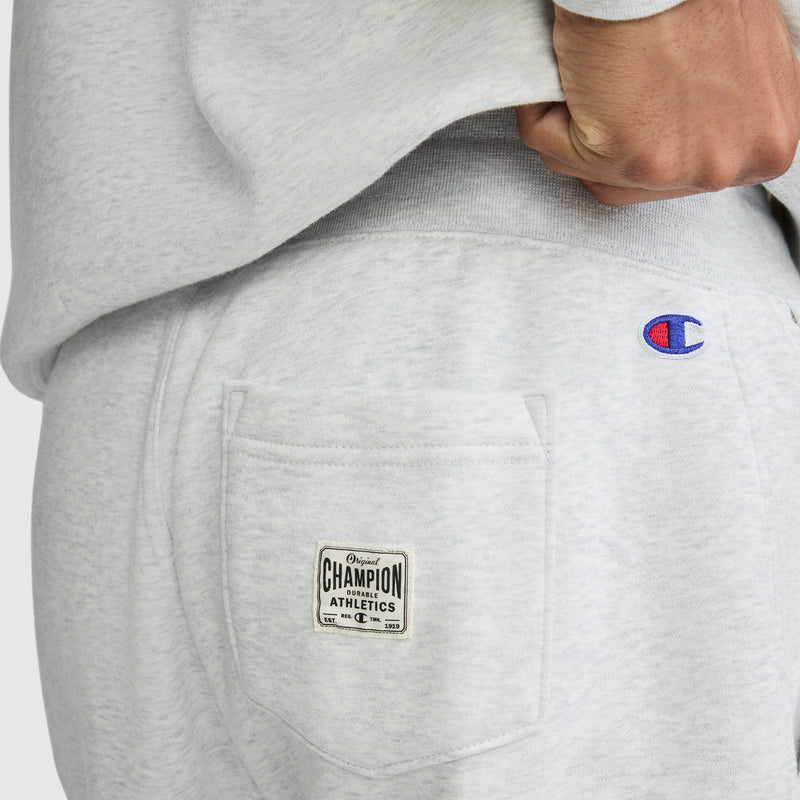 A close-up showcases a person wearing light gray Champion All in a Day’s Work Sweatpants. The photo highlights the back pocket with its Champion Athletics label and embroidered Champion logo, as a hand rests near the waistband—perfect fleece sweatpants for an easygoing, workwear-inspired look.