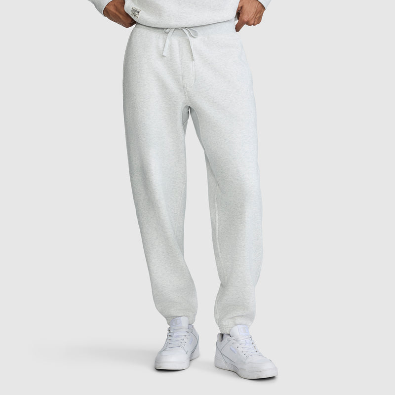 A person wearing Champion All in a Day's Work Sweatpants in light gray with a drawstring, paired with white sneakers. The individual stands with hands at their waist, and the upper body is partially visible, showing a matching sweatshirt. The background is plain white.