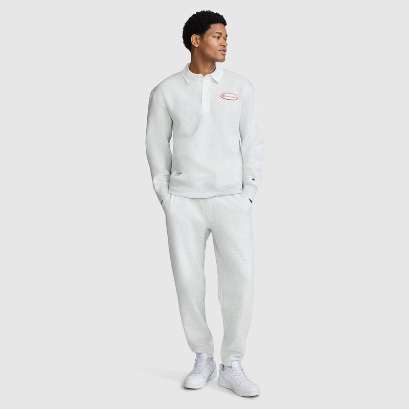A person stands wearing a light gray long-sleeve collared shirt and the Champion All in a Day's Work Sweatpants, paired with white sneakers. The shirt features a small red and white patch on the chest.