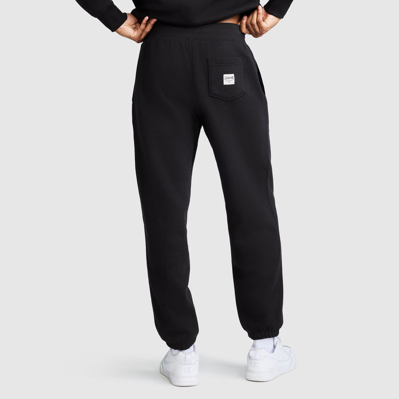 A person stands with hands on hips, wearing Champion's All in a Day's Work Sweatpants in black, paired with a black sweatshirt and white sneakers. The sweatpants feature a back pocket adorned with a white label. The background is plain and light-colored.