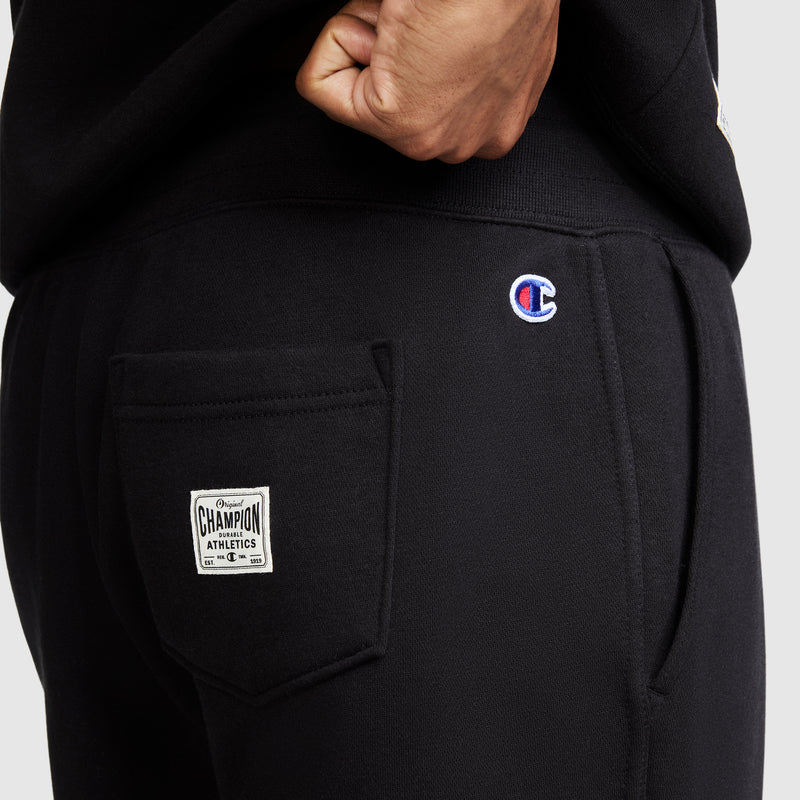 A close-up of a person wearing black "All in a Day's Work Sweatpants" by Champion, featuring a patch on the back pocket and the Champion logo embroidered above it. These athletic fleece pants combine comfort and style for everyday wear.