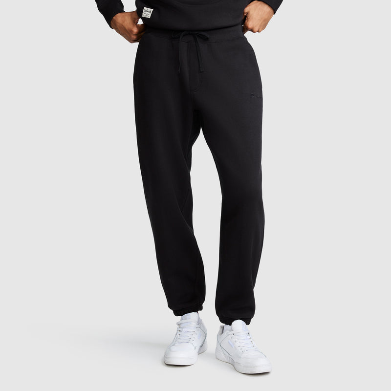A person is shown from the waist down, standing against a plain light gray background, wearing Champion’s All in a Day's Work Sweatpants in black and white sneakers. Their hands rest casually in or near the pockets of the fleece pants.