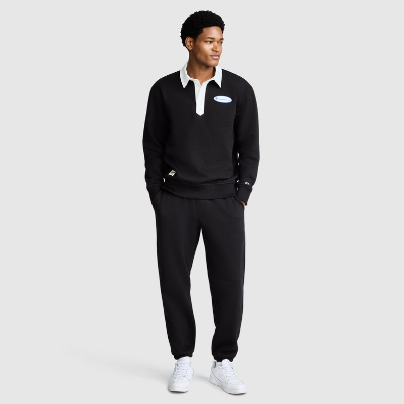 A man stands facing slightly left, wearing a black collared sweatshirt with a white patch, All in a Day's Work Sweatpants by Champion in black, and white sneakers, against a plain light gray background.