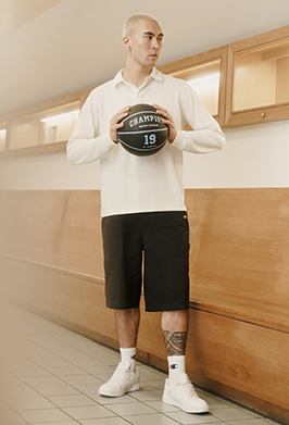 A man stands in a hallway holding a black basketball with 'CHAMPION' and the number 19 printed on it. He is wearing a cream long-sleeve polo shirt, black shorts, white socks, and white sneakers.