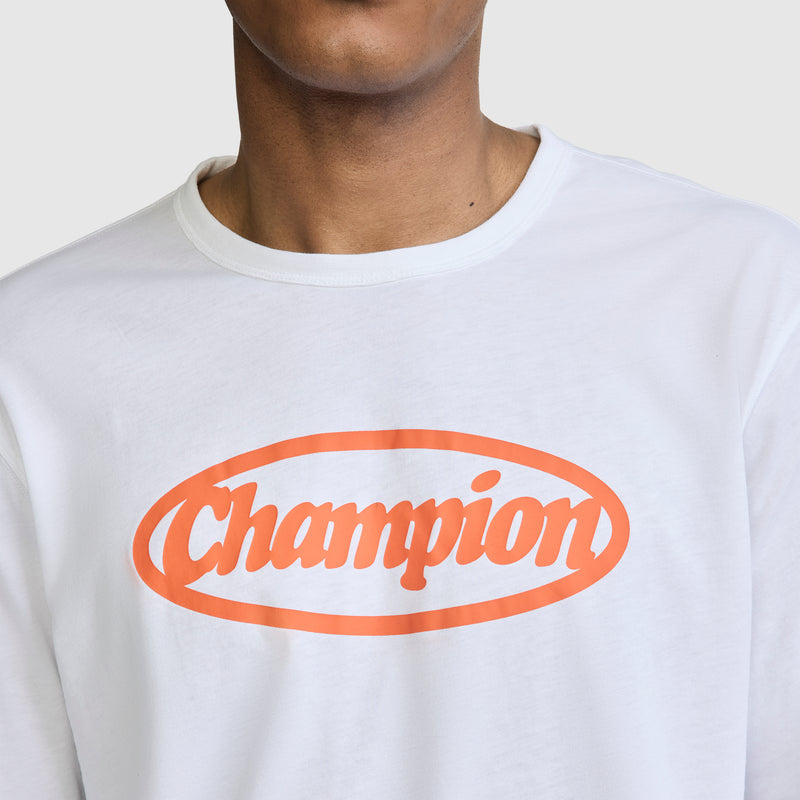 A person wearing the Champion Medallion Long Sleeve Graphic T-Shirt, featuring the brand name in bold orange cursive letters inside an orange oval. The image focuses on the upper torso and neck against a plain background, highlighting this motorsport-inspired shirt.