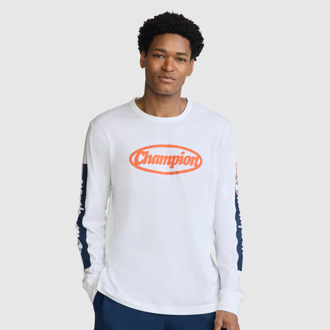 A man wearing the Champion Medallion Long Sleeve Graphic T-Shirt, which features an orange oval medallion logo on the chest and blue panels on the sleeves for a motorsport-inspired look, stands against a plain light background.