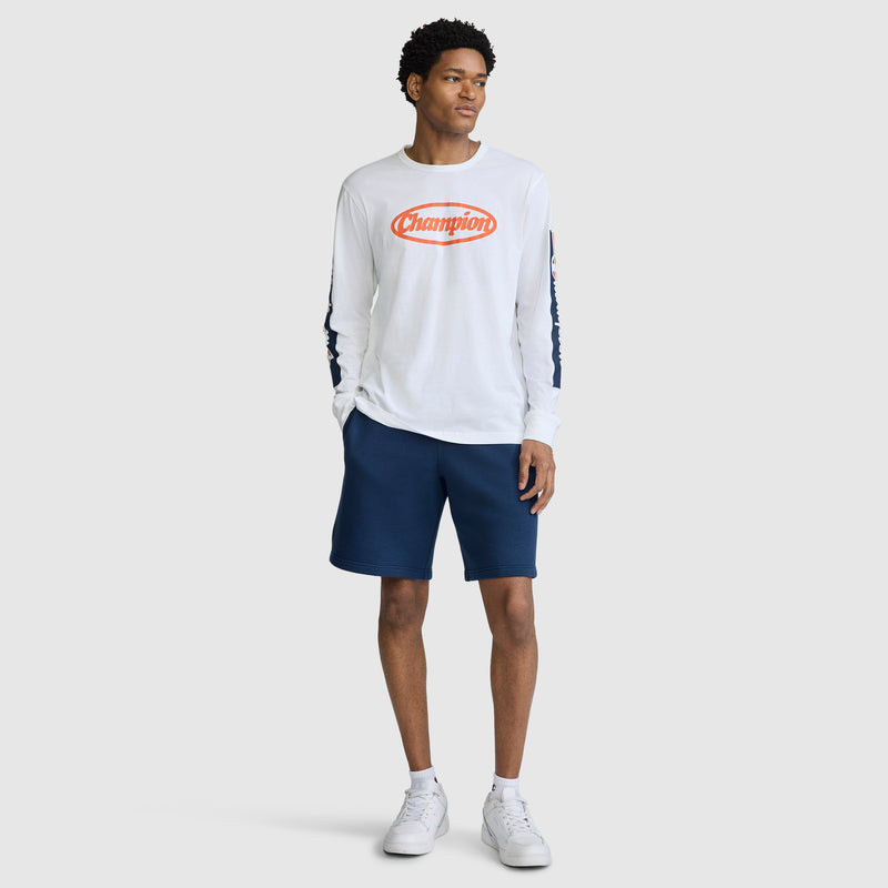 Wearing the Champion Medallion Long Sleeve Graphic T-Shirt in white, paired with navy blue shorts and white sneakers, a person stands against a plain light gray background.