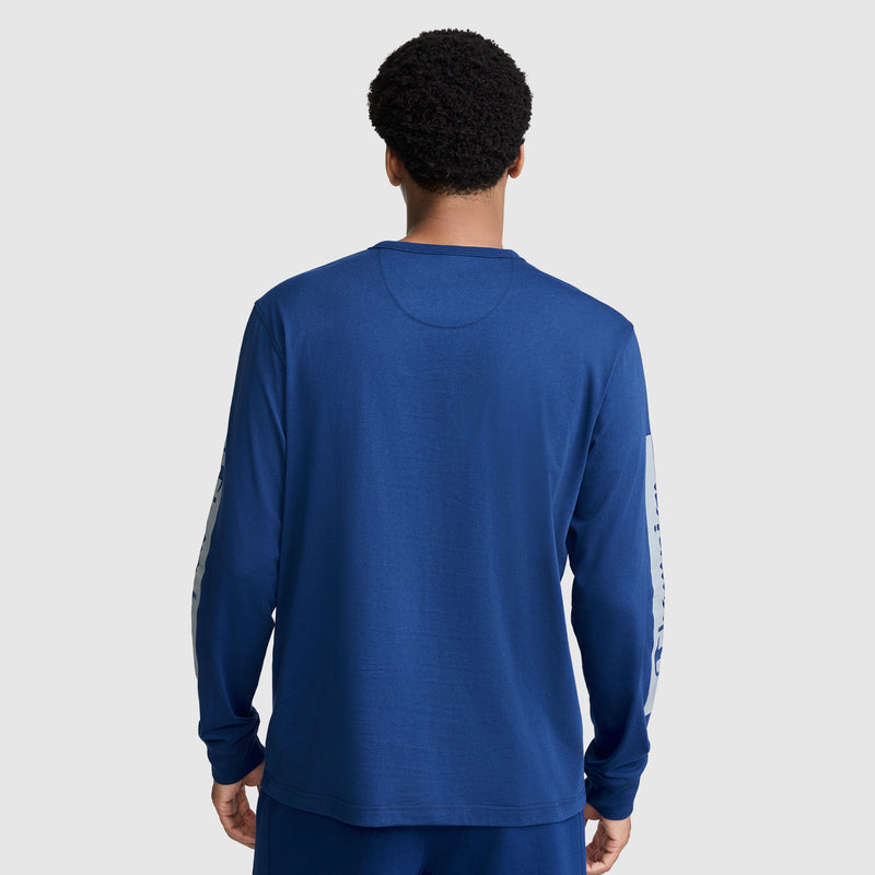 A person stands facing away, wearing a Champion Medallion Long Sleeve Graphic T-Shirt in blue with matching blue pants. The shirt features white text running down the sleeves, reflecting moto sport heritage. The background is plain light gray.