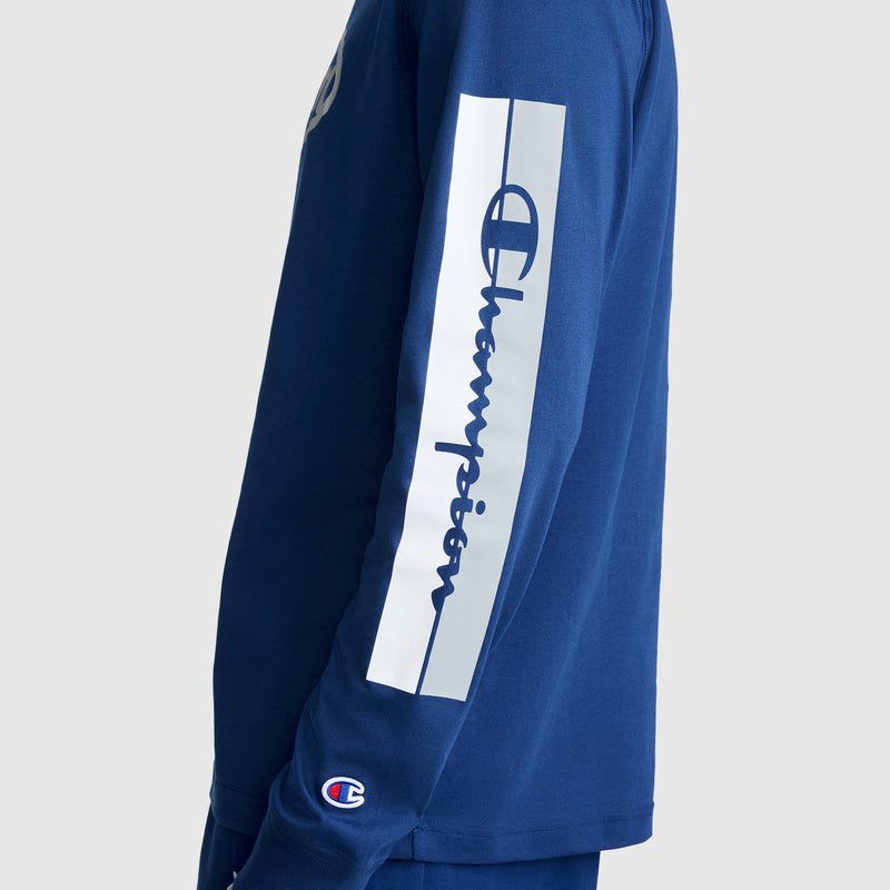 A person wears the Champion Medallion Long Sleeve Graphic T-Shirt in blue, featuring bold “Champion” script along the sleeve and the signature Champion logo on the cuff. Inspired by moto sport heritage, this men’s graphic tee is viewed from the side to highlight its unique sleeve details.