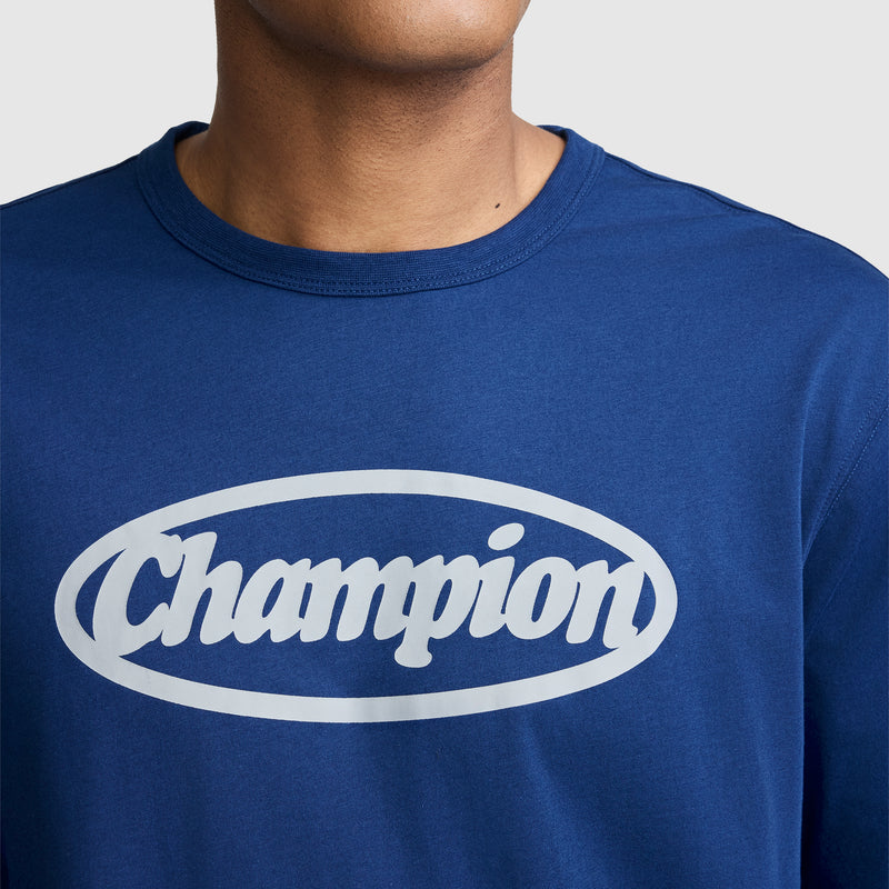 A person wearing a blue Champion Medallion Long Sleeve Graphic T-Shirt, featuring the word “Champion” in white inside an oval on the chest. Only the lower half of their face and neck are visible, subtly referencing moto sport heritage style.