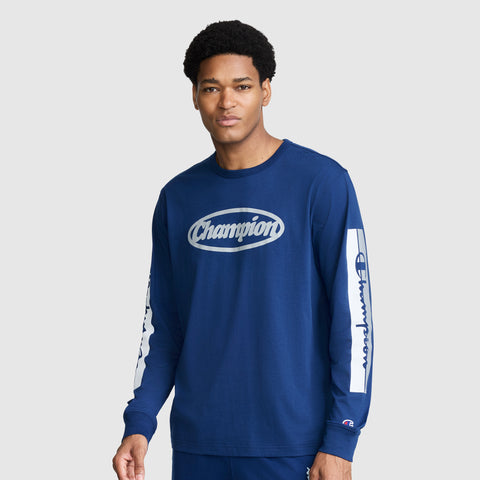 A man wearing the Champion Medallion Long Sleeve Graphic T-Shirt—a blue long sleeve tee with bold white Champion logos on the chest and sleeves—stands against a plain light gray background, channeling classic moto sport heritage style.