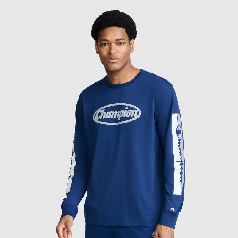 A man wearing the Champion Medallion Long Sleeve Graphic T-Shirt—a blue long sleeve tee with bold white Champion logos on the chest and sleeves—stands against a plain light gray background, channeling classic moto sport heritage style.