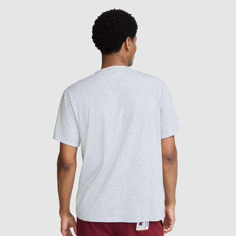 A person with short curly hair, wearing the Champion NY First Squad Graphic T-Shirt in light gray and maroon shorts, is shown from behind against a plain light background, subtly evoking athletic heritage associated with the Champion brand.