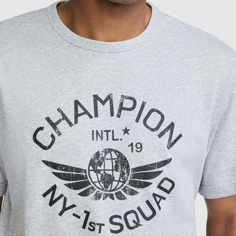 A person wearing a light gray Champion NY First Squad Graphic T-Shirt by Champion, featuring a black CHAMPION INTL. 19 NY-1st SQUAD print and a winged globe graphic on the front, celebrating athletic heritage.