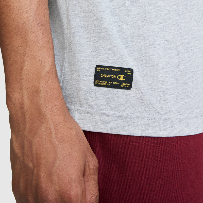 A close-up of a person wearing the Champion NY First Squad Graphic T-Shirt in light gray, featuring the iconic Champion label near the hem and paired with red pants. Only part of the torso and one arm are visible, highlighting Champion’s athletic heritage.