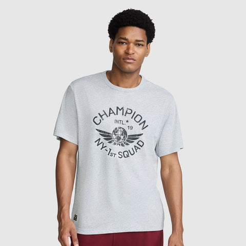 A man wearing the Champion NY First Squad Graphic T-Shirt by Champion, featuring “CHAMPION INTL 19 NY-1ST SQUAD” and a winged globe graphic in light gray, stands against a plain light background, representing the athletic heritage of the NY 1st Squad.
