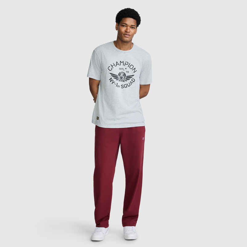 A man stands against a plain background, hands behind his back and wearing a relaxed expression, dressed in a light gray Champion NY First Squad Graphic T-Shirt—a tribute to Champion’s athletic heritage—paired with maroon sweatpants and white sneakers.