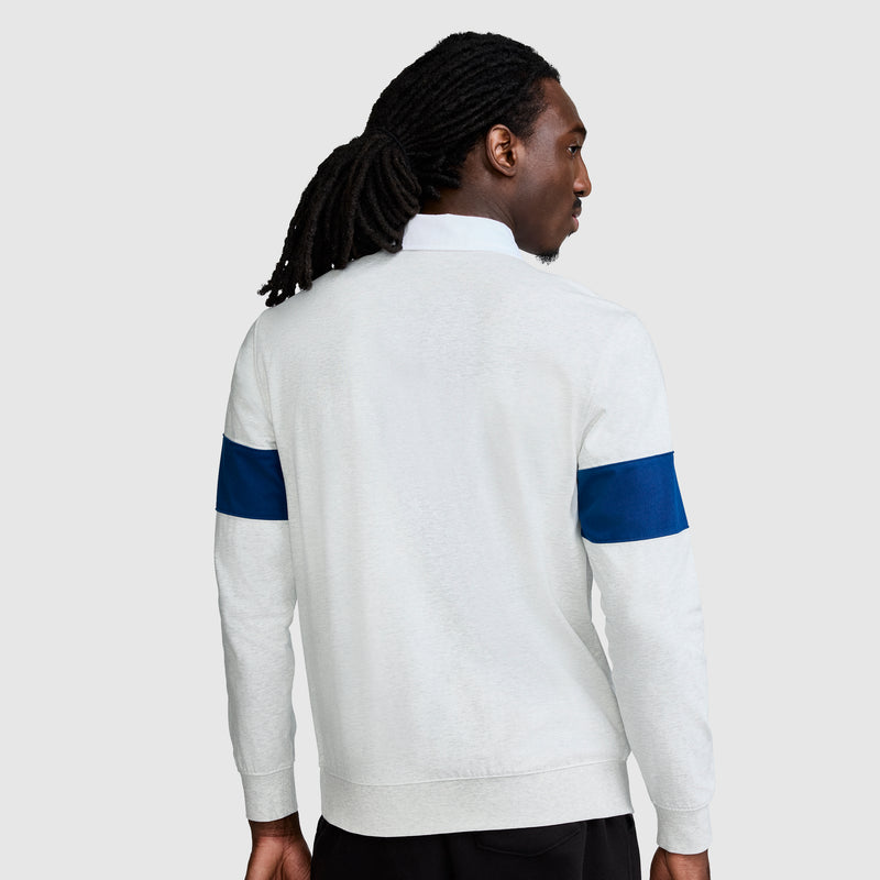 A person with long, dark hair pulled back is seen from behind, wearing the Champion Striped Rugby Shirt—an athletic-style, long-sleeve shirt featuring dark blue sleeve stripes—while standing against a plain light gray background.