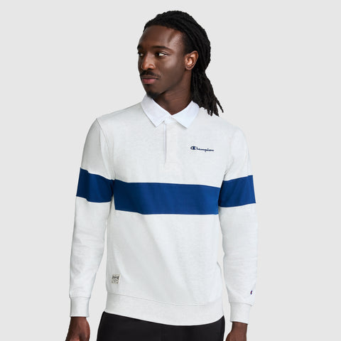 A man with long dark hair models the Champion Striped Rugby Shirt, a white long sleeve shirt designed with a bold blue horizontal stripe and the signature Champion logo, standing against a simple gray background that accentuates its sporty look.