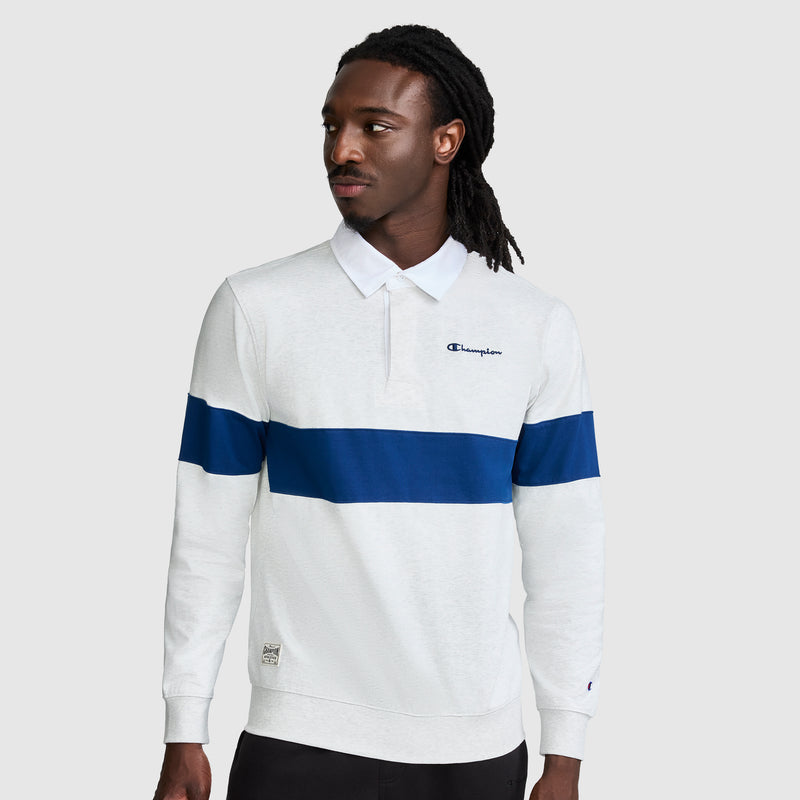 A man with long dark hair models the Champion Striped Rugby Shirt, a white long sleeve shirt designed with a bold blue horizontal stripe and the signature Champion logo, standing against a simple gray background that accentuates its sporty look.