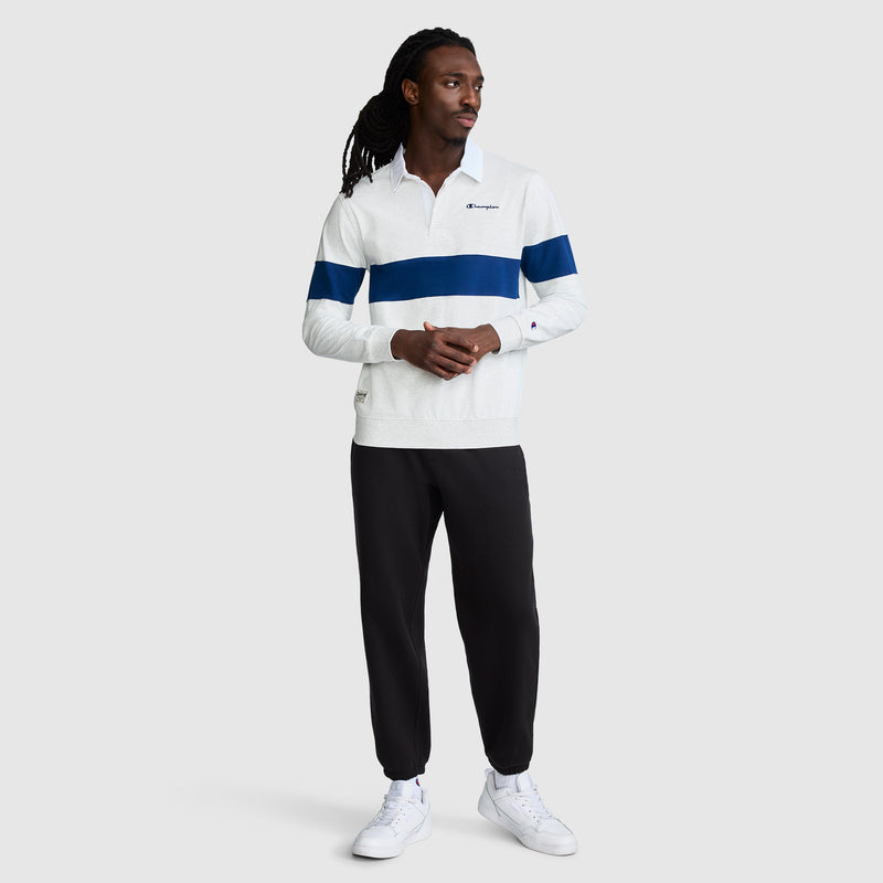 A man stands in a relaxed pose, highlighting an athletic look in the Champion Striped Rugby Shirt—white with a blue stripe—styled with black jogger pants and white sneakers against a plain light gray background.