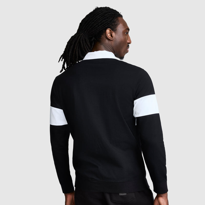A person with long, dark hair pulled back is shown from the back against a light gray background, wearing the Champion Striped Rugby Shirt—a black long-sleeve style with white stripes on the sleeves—capturing an effortless athletic look.