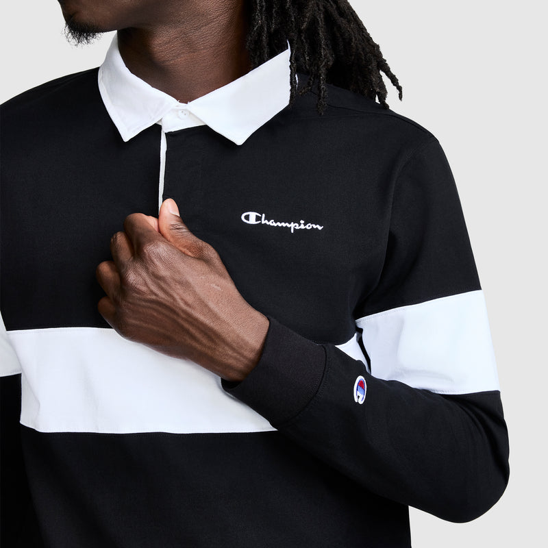 A person wearing the Champion Striped Rugby Shirt—a long sleeve black and white rugby shirt with a sharp white collar—holds the shirt near their chest. The Champion logo appears on both the chest and sleeve, showcasing a timeless athletic look.