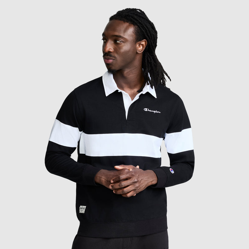 A man with long dreadlocks wears the Champion Striped Rugby Shirt—a black and white, collared long-sleeve shirt—standing against a plain gray background, looking to his left with his hands together, exuding an athletic style.
