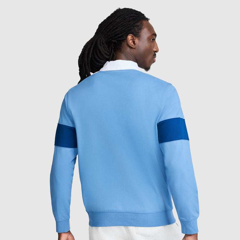 A person with long, dark hair in a ponytail is wearing the Champion Striped Rugby Shirt, featuring an athletic style in light blue with dark blue bands on the sleeves, standing with their back to the camera against a plain light gray background.