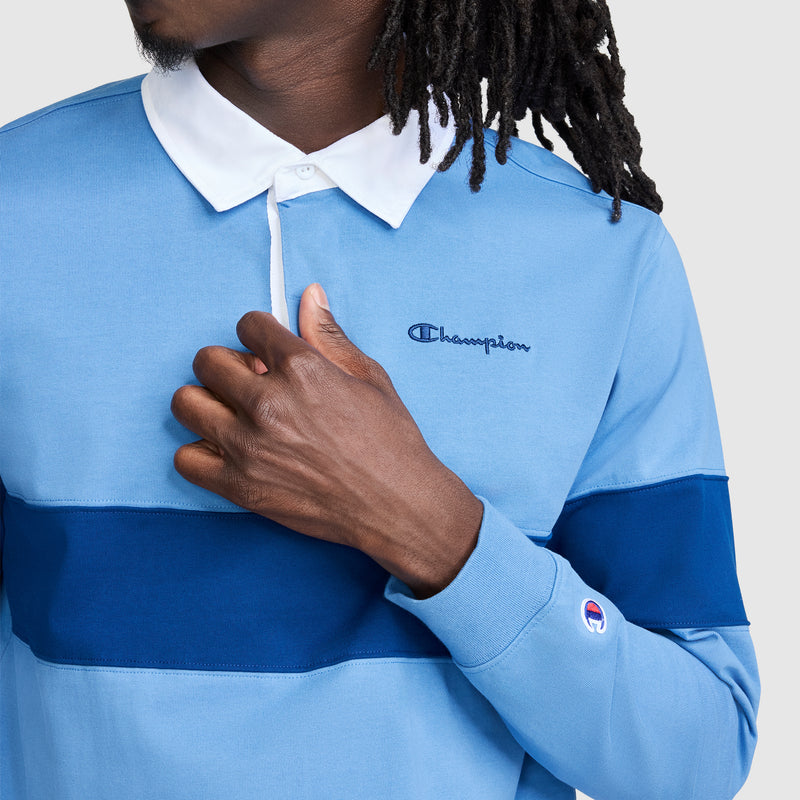 A person with long dreadlocks wears a light blue Champion Striped Rugby Shirt featuring a dark blue stripe and white collar, touching the long sleeve shirt near the Champion logo embroidered on the chest.