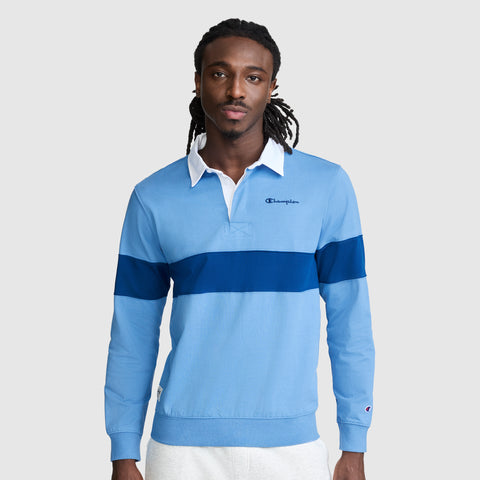 A man stands facing forward wearing a Champion Striped Rugby Shirt in light blue, featuring a dark blue stripe and a white collar. The long sleeve shirt showcases an athletic style with the Champion logo on the chest. The background is plain light gray.