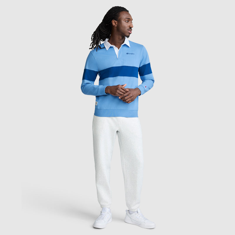 A man with long hair is seen wearing the Champion Striped Rugby Shirt in light blue with dark blue stripes, styled with white pants and sneakers for a laid-back athletic look against a simple light background.