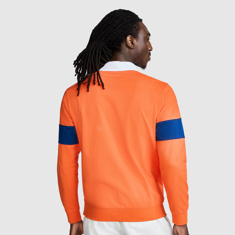 A person with long braided hair is seen from behind against a plain light background, wearing the Champion Striped Rugby Shirt layered under an orange jacket with a blue stripe on the sleeve.