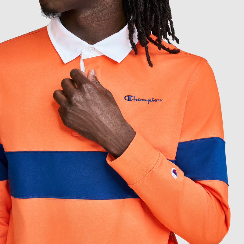 A person with dark skin and dreadlocks wears the Champion Striped Rugby Shirt, featuring bold orange and blue color-blocking and a white collar. They are adjusting the button placket on this men's shirt, which displays the Champion logo embroidered on both the chest and sleeve cuff.