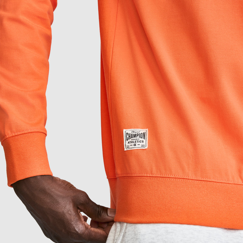 A person wearing an orange Champion Athletics sweatshirt and light gray pants, with a close-up on the Champion label near the hem and their left hand visible—perfect for pairing with the Champion Striped Rugby Shirt for a sporty look.