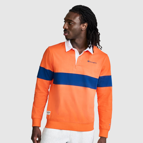 A man with long hair stands against a plain light background, looking to his right, wearing the Champion Striped Rugby Shirt in orange with a blue horizontal stripe and white collar, paired with white pants.
