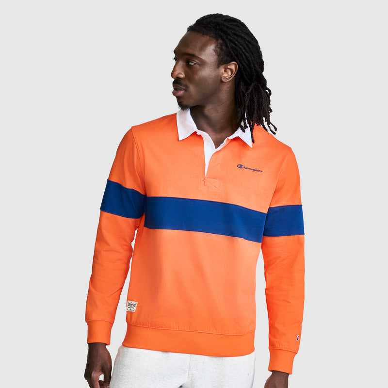 A man with long hair stands against a plain light background, looking to his right, wearing the Champion Striped Rugby Shirt in orange with a blue horizontal stripe and white collar, paired with white pants.
