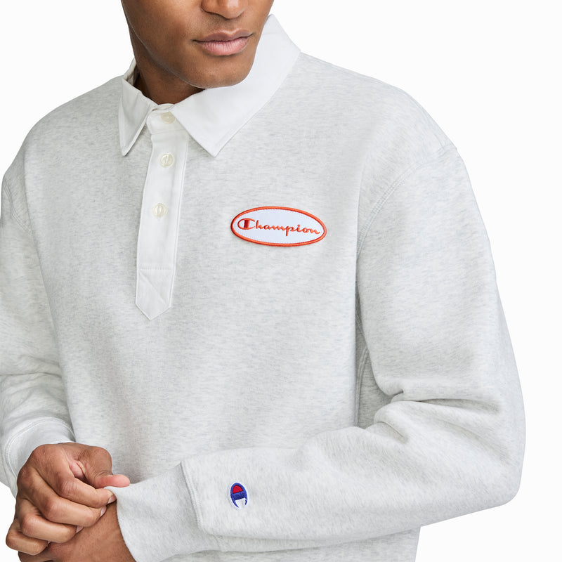 A person is wearing the Champion Fleece Rugby, a men’s rugby shirt in light gray featuring a white collar and red embroidered Champion logo on the chest. The Champion logo is also visible near the left sleeve cuff. The person's face is partially out of frame.