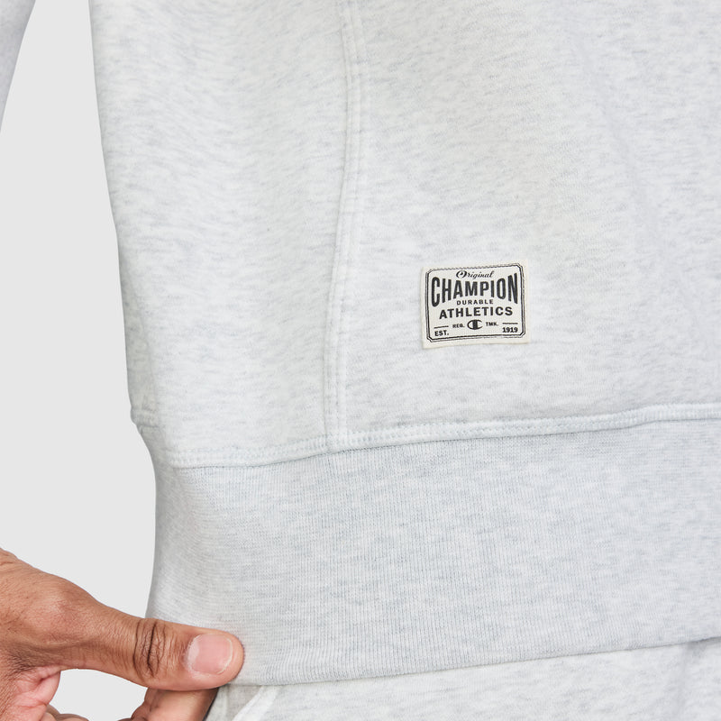 A close-up of a person wearing the Champion Fleece Rugby in light grey, featuring a small logo patch near the hem. The person is holding the hem of the Fleece Rugby with their left hand.