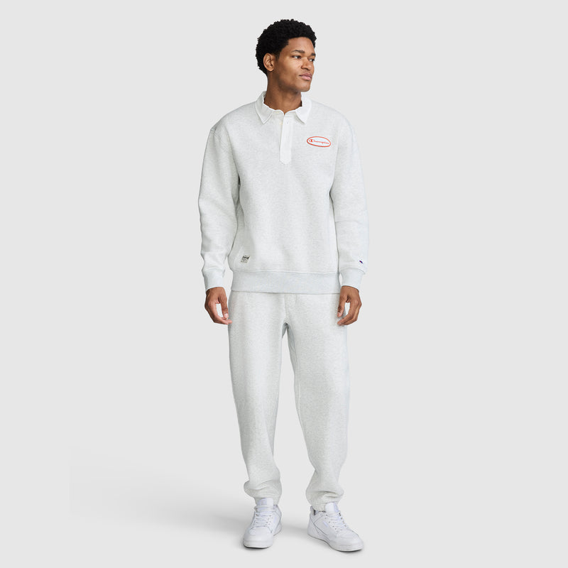 A person stands wearing a light gray Champion Fleece Rugby pullover and matching sweatpants with white sneakers. The Champion Fleece Rugby features a collar, buttons, and a red Champion logo on the chest. The background is plain and light.